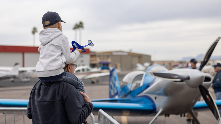 Young boy on top of his dad's shoulders, looking at a plane while holding a paper airplane