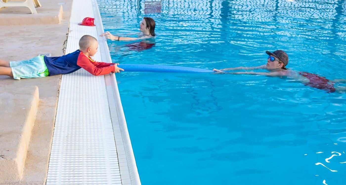 Child laying on a pool deck holding a blue pool noodle to an adult in the pool
