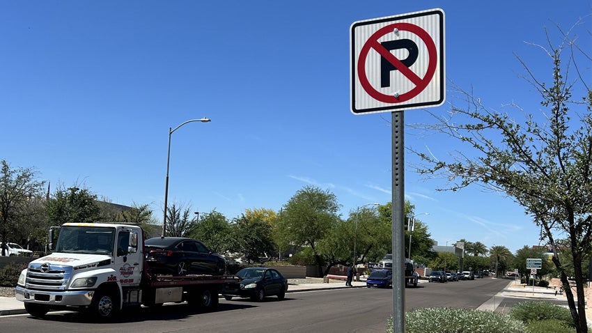 No parking sign on a street with a vehicle on a tow truck in the background.