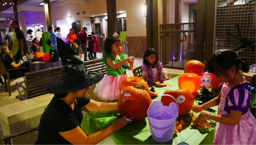 young girls dressed in princess costumes, carve pumpkins outside on a table