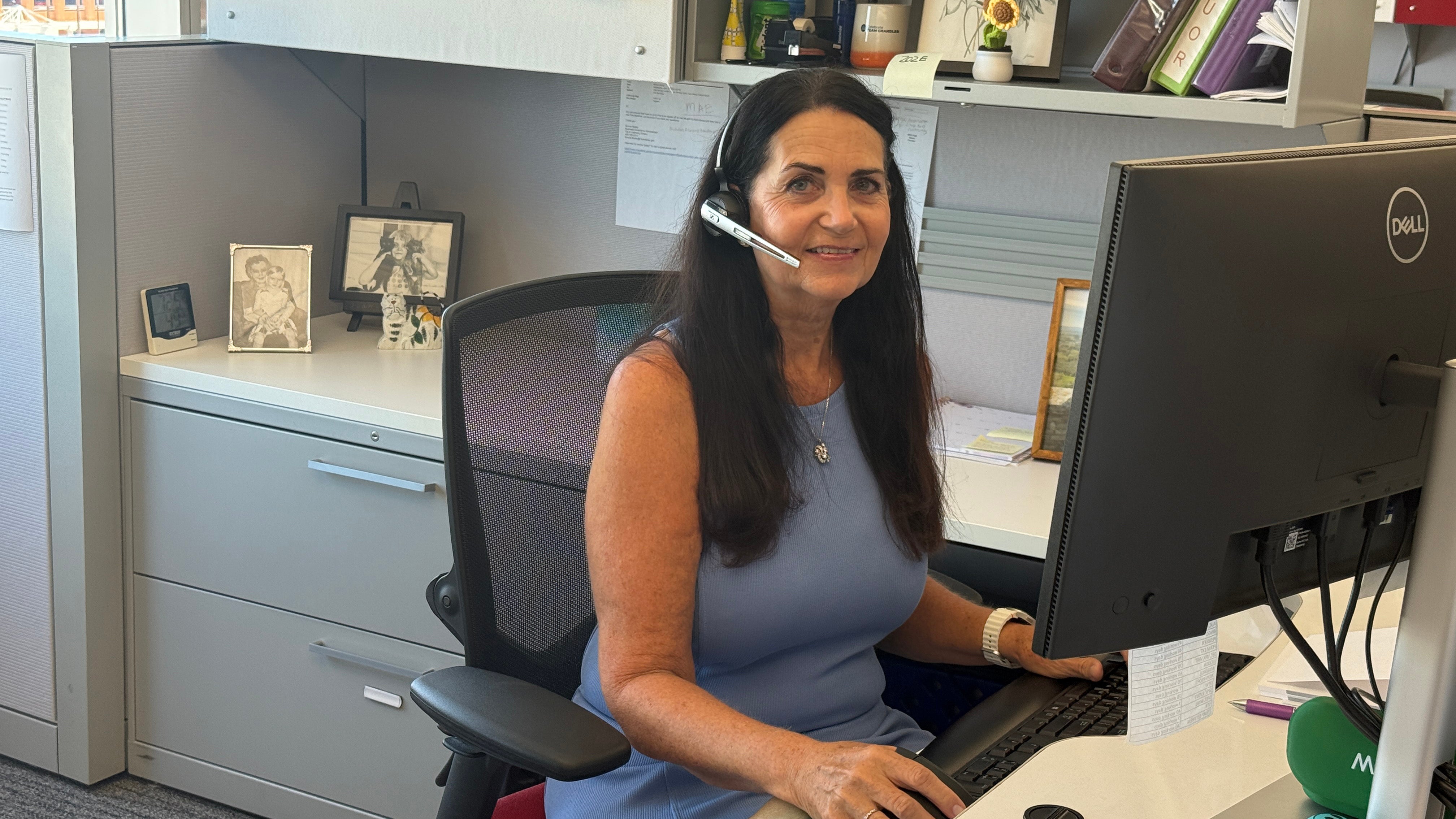 A woman in a light blue dress is sitting down at her computer smiling at the camera while taking a phone call