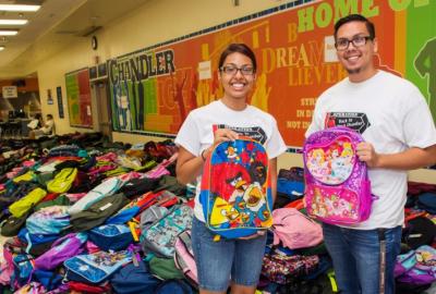 Two volunteers holding backpacks in front of a large pile of backpacks