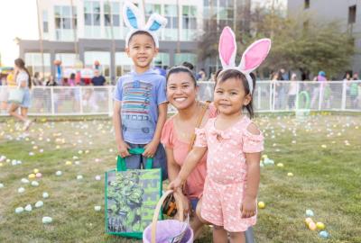 Young boy, woman and young girl wearing bunny ears crouching in front of a grassy field filled with colorful Easter eggs