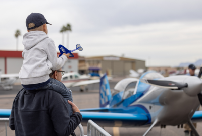 Little boy on his parent's shoulder holding a paper plane while looking at a plane