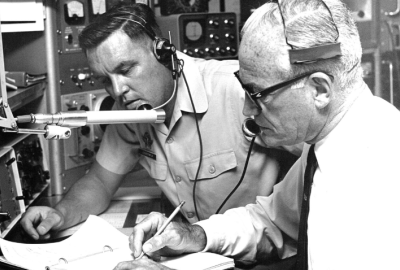 Two men with headsets on looking at a sheet of paper in a radio station