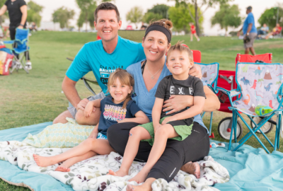 A family of four sits together on a blue picnic blanket spread across a grassy field at Tumbleweed Park. A man in a bright blue t-shirt and a woman with a dark headband and blue shirt are smiling at the camera. Their young children are sitting in the woman's lap. The girl, wearing a dark blue shirt, and the boy, in a grey t-shirt and green shorts, are also smiling brightly. In the background, other people are visible on the lawn, along with folding chairs, under the soft light of late afternoon.