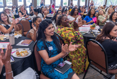 A large group of diverse women sit at round banquet tables, smiling and clapping enthusiastically