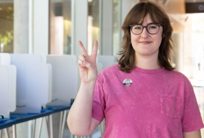 Girl wearing a pink t-shirt holding up a peace sign with an "I Voted" sticker on her shirt. She's standing in front of a row of voting booths.