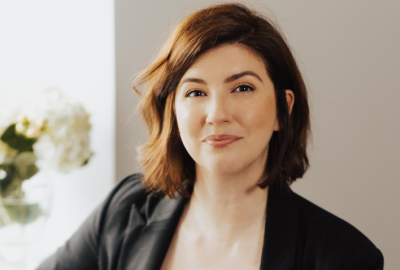 Woman with short brown hair wearing a black blazer is smiling at the camera with a white wall behind her