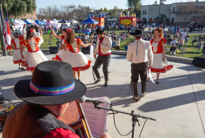 Male and female Folklórico dancers on an outdoor stage with the audience and a festival seen beyond them