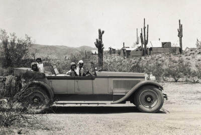 Black and white photo of a couple in an old convertible in the middle of the desert with cabins behind them