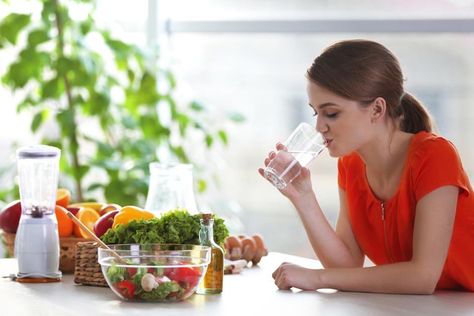 Young woman drinking water surrounded by veggies on the counter
