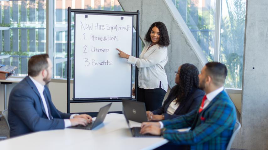 A group of employees sitting at a training table for a meeting talking about items listed on a whiteboard