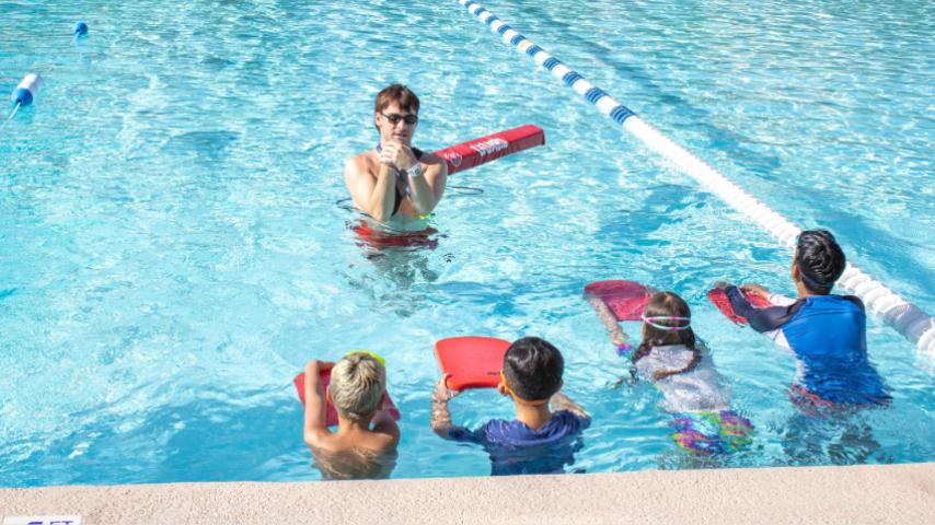 four kids with their backs to a wall in a swimming pool. Looking at the swim instructor showing a stroke technique. 