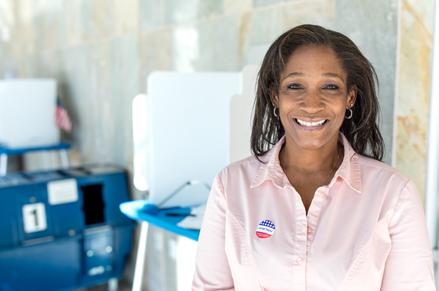 Smiling female voter at election site with an I Voted sticker on her pink shirt with election booth in the background