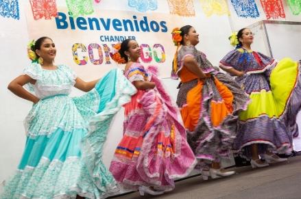Four ballet folklorico dancers wearing coloring dresses dancing in front of a sign that reads Bienvenidos Chander Contigo