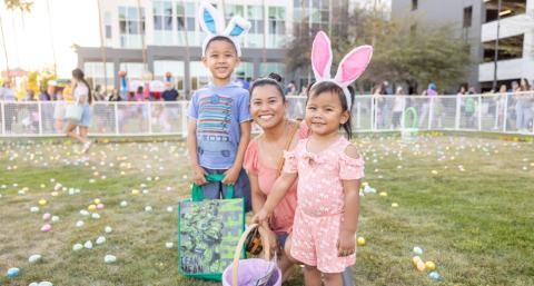 Young boy, woman and young girl wearing bunny ears crouching in front of a grassy field filled with colorful Easter eggs