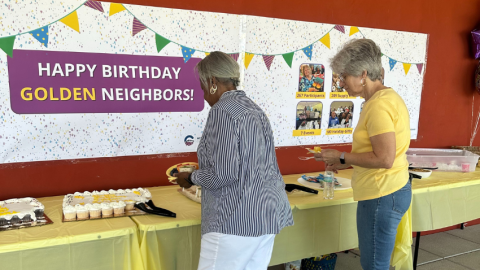 Two female senior residents getting cake with a "Happy Birthday Golden Neighbors" banner in the background