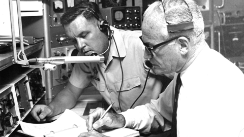 Two men with headsets on looking at a sheet of paper in a radio station