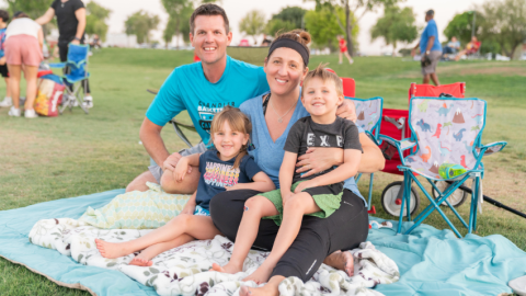 A family of four sits together on a blue picnic blanket spread across a grassy field at Tumbleweed Park. A man in a bright blue t-shirt and a woman with a dark headband and blue shirt are smiling at the camera. Their young children are sitting in the woman's lap. The girl, wearing a dark blue shirt, and the boy, in a grey t-shirt and green shorts, are also smiling brightly. In the background, other people are visible on the lawn, along with folding chairs, under the soft light of late afternoon.