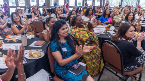 A large group of diverse women sit at round banquet tables, smiling and clapping enthusiastically