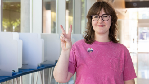Girl wearing a pink t-shirt holding up a peace sign with an "I Voted" sticker on her shirt. She's standing in front of a row of voting booths.