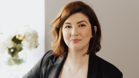 Woman with short brown hair wearing a black blazer is smiling at the camera with a white wall behind her