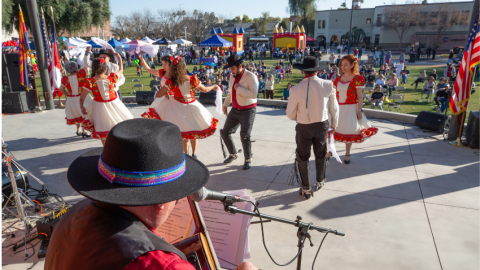Male and female Folklórico dancers on an outdoor stage with the audience and a festival seen beyond them