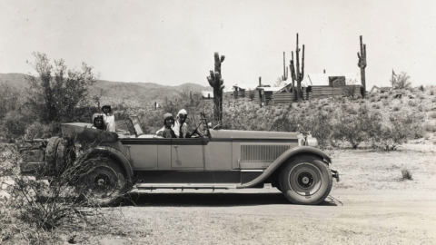 Black and white photo of a couple in an old convertible in the middle of the desert with cabins behind them